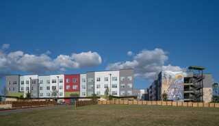 Wide exterior view of the Norman Commons apartment community on a clear day, featuring a modern four-story residential building with white, gray and red façade panels, large windows and green accents. A second building connected by a central walkway displays a large colorful mural on its end wall. A grassy open space and wood fencing frame the development beneath a bright blue sky with scattered clouds.