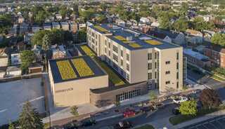 A modern multi-story building features green rooftop gardens and is surrounded by residential areas and streets, showcasing urban development.