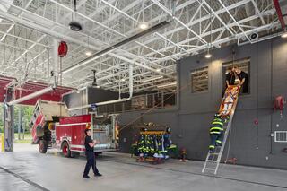 Firefighters practice rescue techniques using a ladder and harness, with a fire truck parked nearby in a well-lit station.