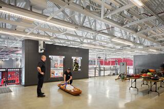 Two firefighters are in a spacious training facility, one preparing a stretcher while the other observes, surrounded by equipment and tools.