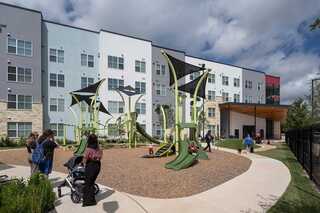 A vibrant playground with unique green structures is surrounded by a modern apartment building, featuring people and strollers in the area.