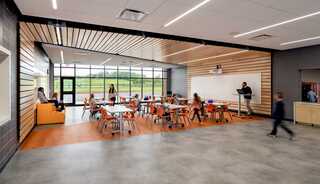 A modern classroom with vibrant orange chairs, children engaged in activities, and large windows allowing natural light and outdoor views.