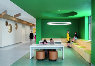 A vibrant, modern space featuring green walls, a large light fixture, and children engaged in activities at a communal table and seating area.