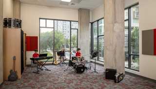 Three children are rehearsing music with a keyboard, drums, and guitar in a bright studio featuring large windows and modern decor.