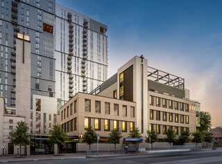 Modern buildings blend sleek glass and contemporary design, with greenery lining the street, evoking an urban, dynamic atmosphere at sunset.