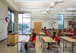 A teacher stands at the front of a modern classroom, while attentive students in red shirts are seated at desks, engaging in learning.