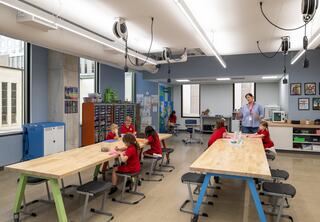 A bright, modern classroom with wooden tables where students in red shirts engage in activities while a teacher supervises.