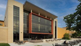 A modern building features large glass windows and a striking red accent, surrounded by outdoor seating and greenery under a blue sky.