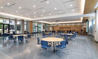 A spacious, modern room features round tables and chairs, large windows, and a projector, with people gathered for an event or meeting.