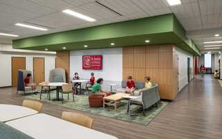 A modern, open educational space features students in casual seating, engaging in discussions, with a vibrant green ceiling and wooden accents.