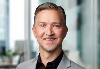 A smiling individual with light gray hair and a neatly trimmed beard wears a gray blazer over a black shirt, set against a blurred office background.