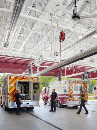 Emergency responders work in a modern fire station, preparing equipment and vehicles in a bright, spacious garage.