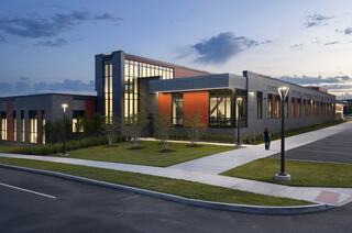 A modern building with a sleek design features large windows and a mix of gray and orange materials, surrounded by landscaped greenery.