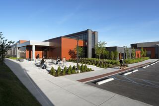 A modern building with orange and gray accents, surrounded by lush landscaping, a bike rack, and a well-maintained parking lot under clear skies.