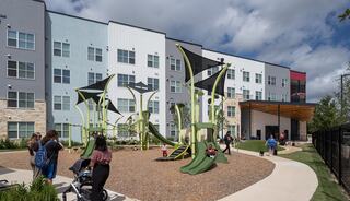 A modern playground features unique green structures, with families and children playing outside a contemporary multi-story building under a cloudy sky.