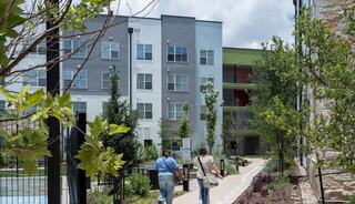Two people walk along a pathway lined with greenery, approaching modern apartment buildings under a partly cloudy sky.