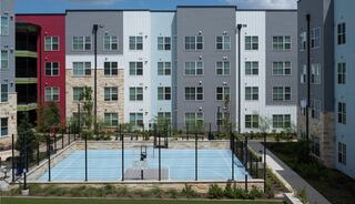 A modern residential complex features colorful buildings surrounding a fenced basketball court with landscaped areas and greenery.