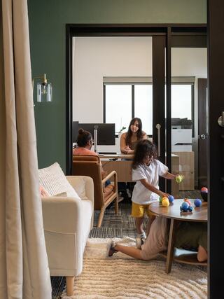 A cozy office space features adults working at desks, while children play on the floor with colorful toys nearby.