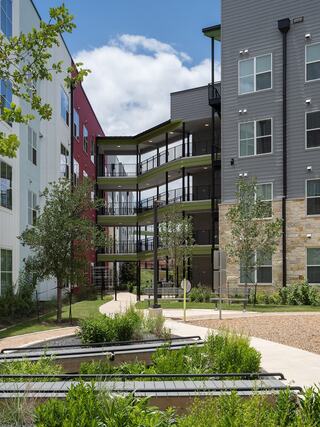 A modern courtyard features two colorful buildings, lush greenery, and a walkway with seating, creating a vibrant community space.