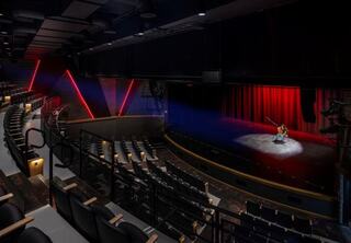 A dimly lit theater featuring an empty audience area, with vibrant red curtains and a lone performer on stage illuminated by focused light.