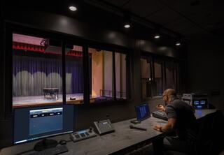 A control room overlooks a stage, with a technician focused on screens and equipment, preparing for an upcoming performance.