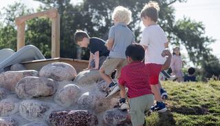 Four children climb a rock formation at a playground, while others play in the background on a sunny day.