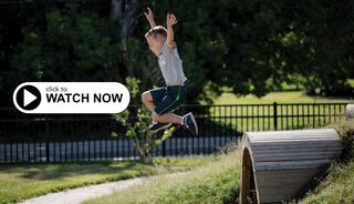 A boy joyfully leaps into the air, playing outdoors in a green, sunny environment, showcasing a moment of excitement and adventure.