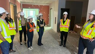 A group of people in hard hats and safety vests listens attentively to a speaker in a construction site with unfinished walls.