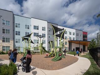 A lively playground with unique green structures sits in front of colorful apartment buildings, as families enjoy the outdoor space.