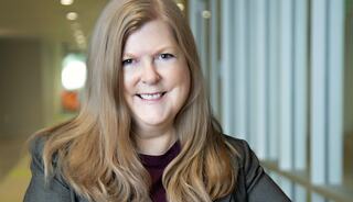 A smiling woman with long hair, wearing a blazer, poses confidently, with soft lighting and a modern backdrop.
