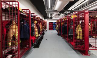 A spacious locker area features rows of red metal racks holding various fire uniforms and equipment, with organized storage for quick access.