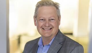 A smiling man in a gray blazer and blue shirt stands confidently, exuding warmth and professionalism in a bright, modern setting.