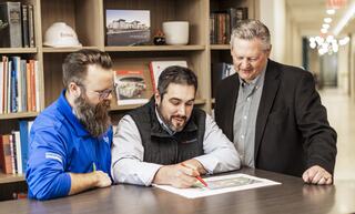 Three professionals collaborate over a plan on a table, discussing and marking details, with bookshelves and a bright office setting in the background.