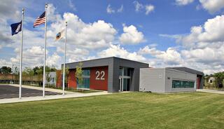 A modern building features large windows, a prominent number 22, and is flanked by flagpoles under a partly cloudy blue sky.