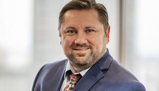A smiling man with a beard wears a blue suit and patterned tie, standing confidently against a blurred office background.