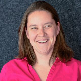 A woman with shoulder-length brown hair smiles warmly, wearing a pink shirt against a dark gray background.