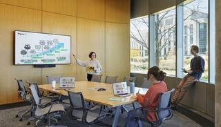 A meeting room features a large screen displaying diagrams. One speaker points at the presentation while others are engaged at their laptops.