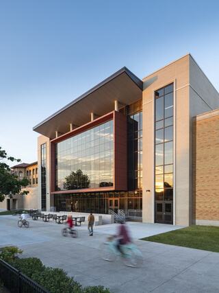 A modern high school entrance with large glass windows and a sleek design, with people biking and walking nearby at sunset.