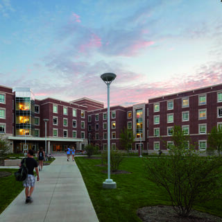 A group of students walk along a pathway lined with greenery and modern brick buildings during a colorful sunset.