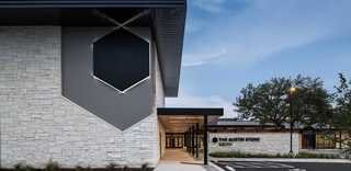 A modern building features a stone facade and sleek design, with a prominent geometric element and inviting entrance under a clear sky.