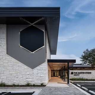 A modern building featuring a stone facade, geometric design elements, and a welcoming entrance, surrounded by greenery and a clear sky.
