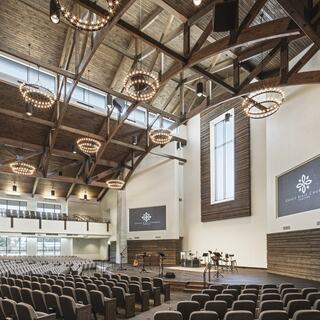 A spacious, modern church interior features high wooden beams, elegant chandeliers, a large window, and a stage with musical instruments.