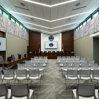 A modern conference room featuring rows of white chairs, a central podium, monitors, and natural light from large windows.