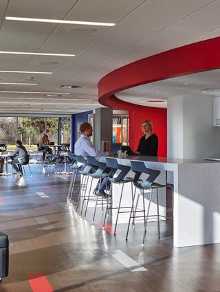 A modern office space featuring a stylish bar area, two people chatting, and others working at tables, infused with natural light.