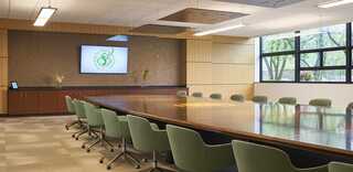 A modern conference room features a long wooden table, green chairs, a wall-mounted screen, and large windows with natural light.