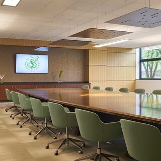 A spacious conference room features a large wooden table, green chairs, a screen displaying a logo, and natural light from a window.