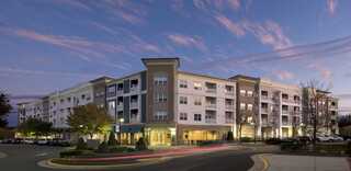 A modern multi-story apartment building stands at dusk, with elegant architecture, landscaped surroundings, and a warm, inviting glow.