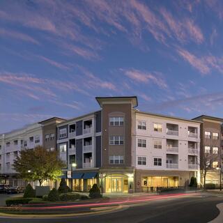 A modern apartment complex with multiple levels, surrounded by trees and parked cars, set against a colorful evening sky.