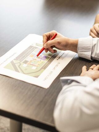 A person points at a colored map on a table while discussing plans, with another hand visible nearby, suggesting collaboration.