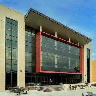 A modern building features expansive glass windows and a bold red accent. Outdoor seating is visible in front, set against a clear sky.